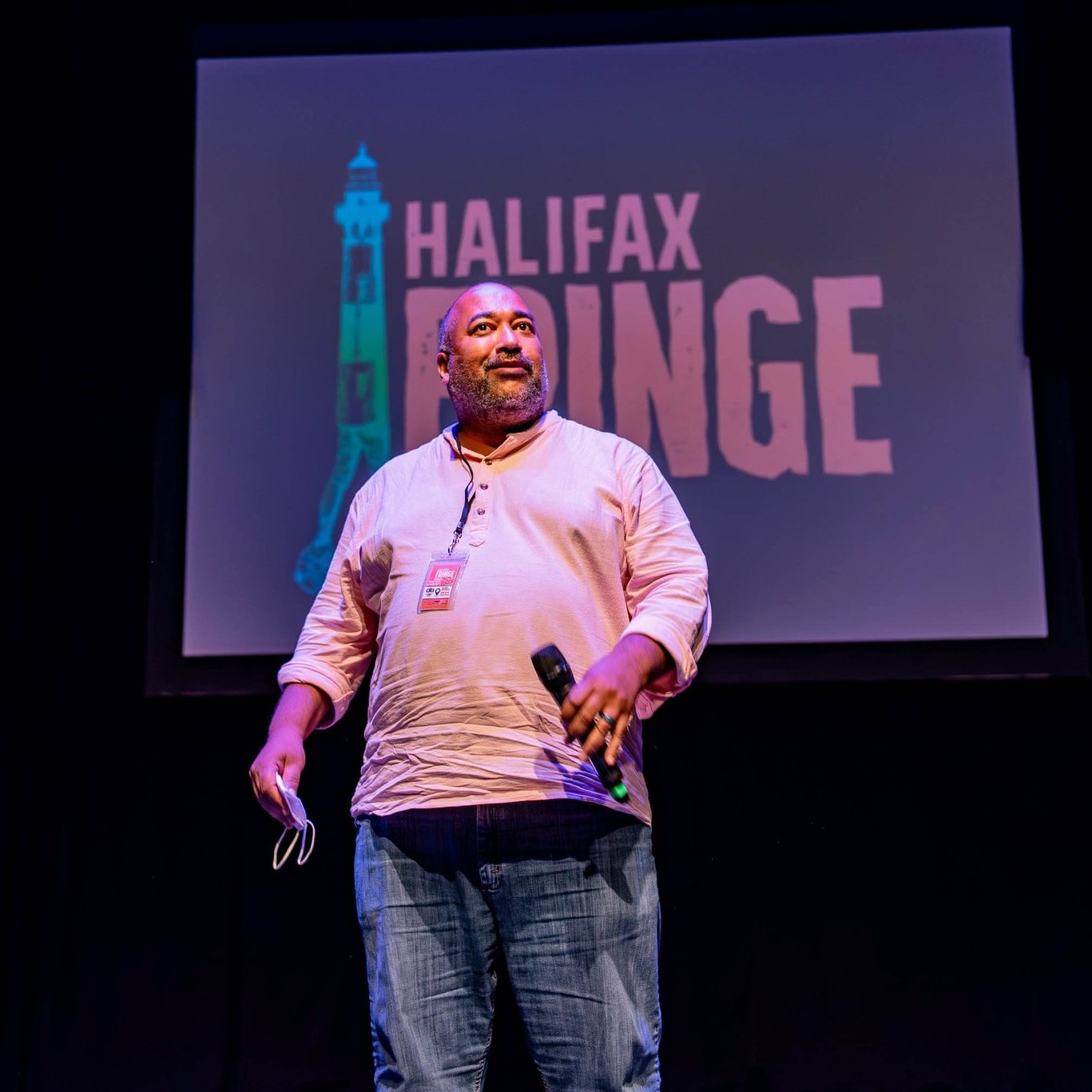 The Velvet Duke stands in front of the Halifax Fringe Festival logo projected on a screen. Velvet is a Black person wearing a light pink shirt and blue jeans. The logo has similar colours but this is surely just coincidence. Credit: Stoo Metz Photography